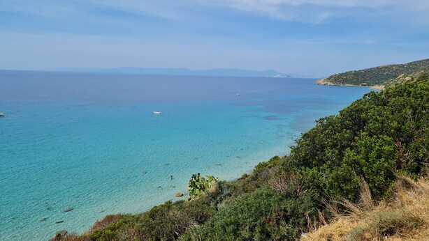 A coastal landscape with clear turquoise waters, a boat in the distance, and dense green vegetation on the right.
