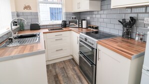 A modern kitchen with white cabinets, a wooden countertop, a built-in oven, a microwave, and a window with blinds. - Sandy Toes Cottage (Newbiggin-by-the-Sea)