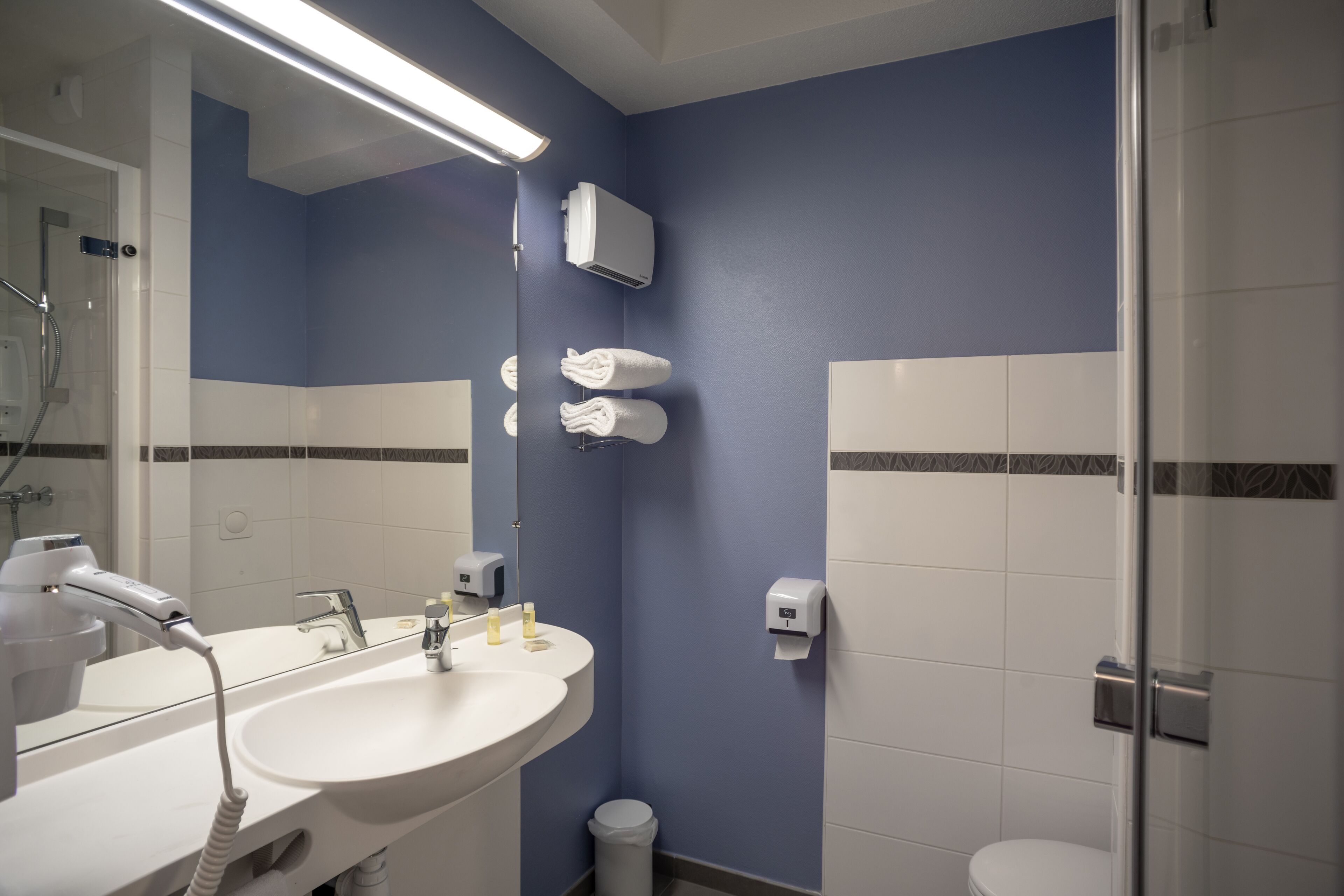 A bathroom with a sink, mirror, towel dispenser, and a glass shower enclosure.