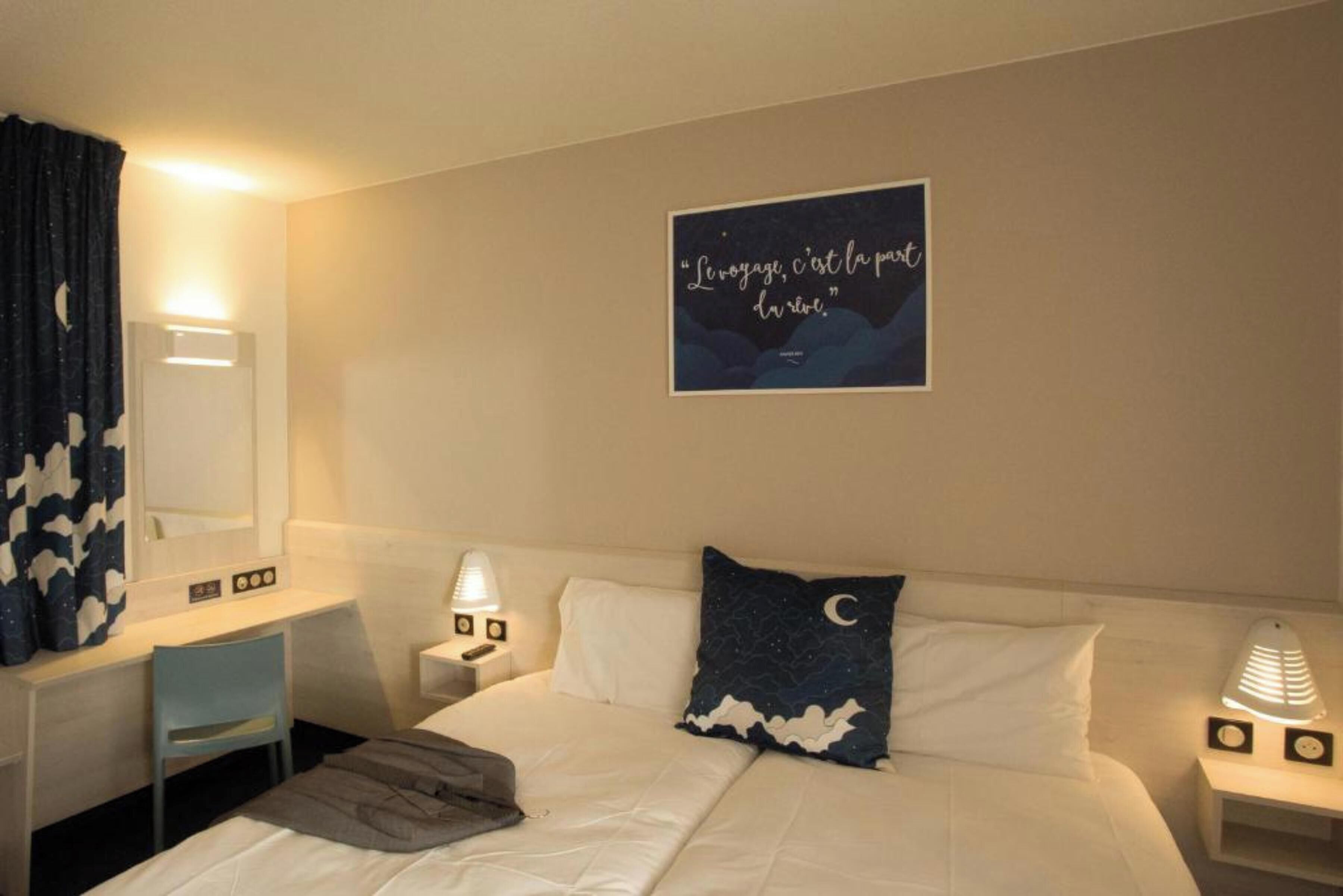 A hotel room with a bed, a desk, a chair, a nightstand with a lamp, and a wall with a framed quote.