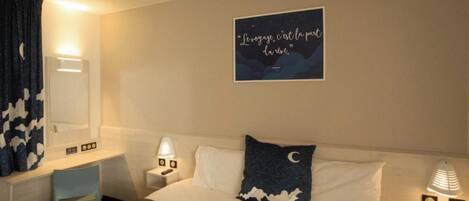 A hotel room with a bed, a desk, a chair, a nightstand with a lamp, and a wall with a framed quote.