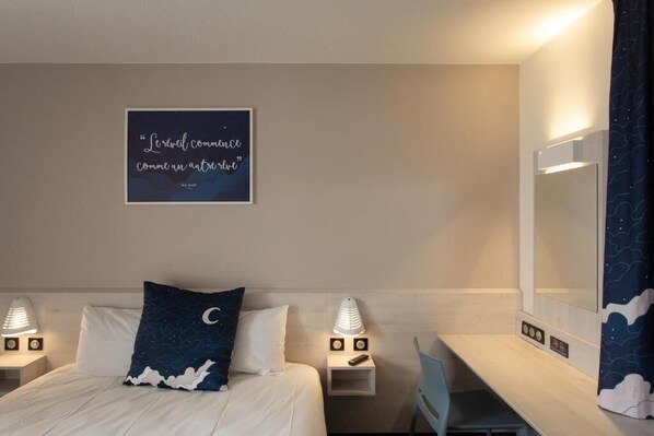 Habitación de hotel con cama, escritorio, mesita de noche, espejo y un cuadro con una cita en la pared.