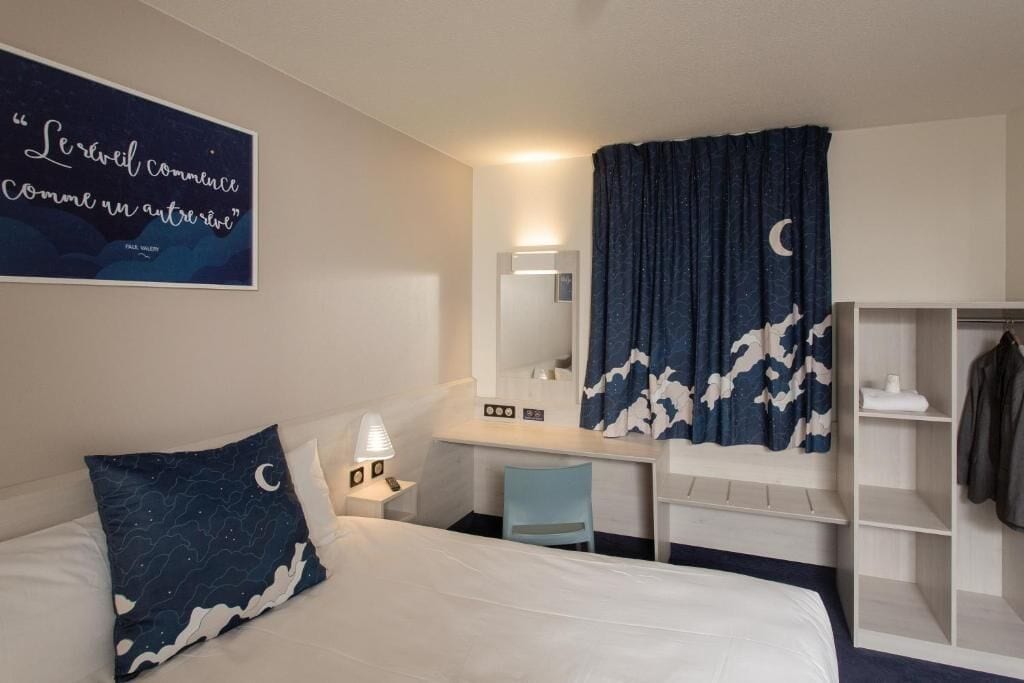 A hotel room with a bed, a desk, a chair, a wardrobe, a mirror, and a wall with a quote.