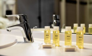 A bathroom sink with a chrome faucet, several yellow bottles labeled 'ace HOTEL' on the countertop, and a mirror reflecting the sink area. - ACE Hôtel Lyon Vénissieux (Venissieux)