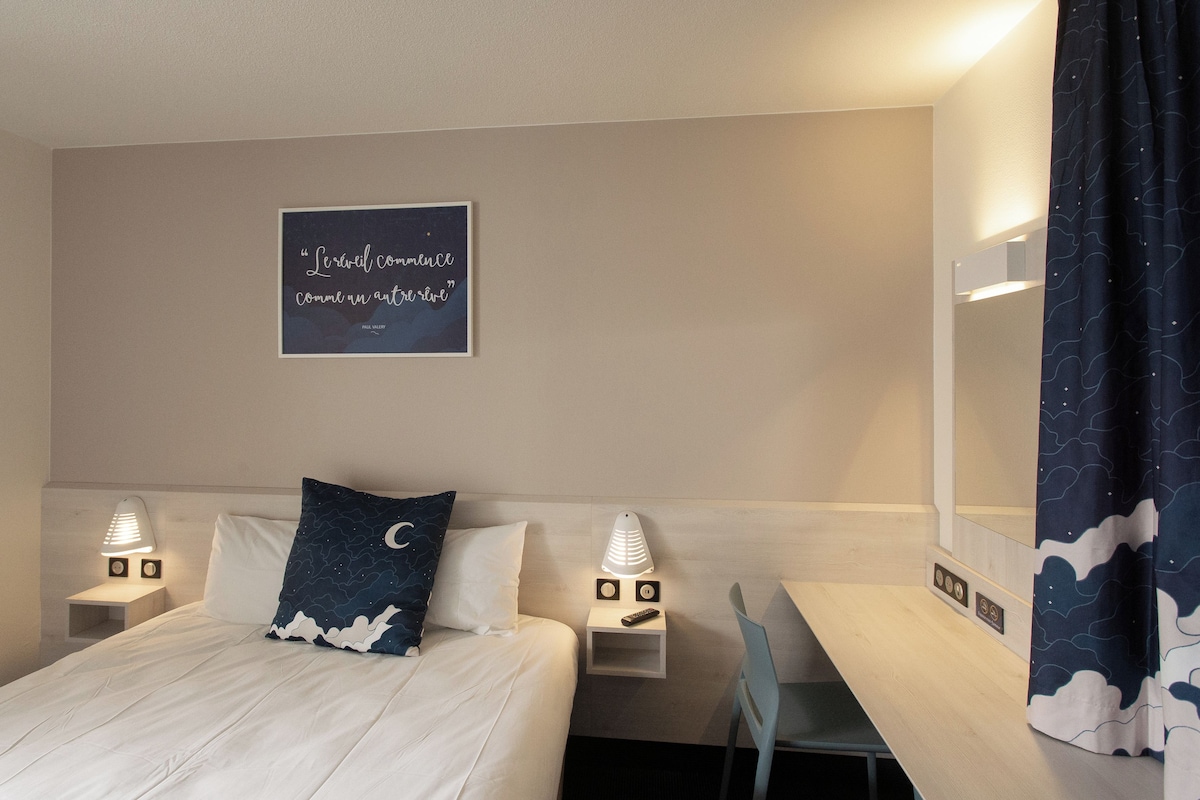 A hotel room with a bed, two bedside tables with lamps, a desk, a chair, and a wall-mounted mirror.