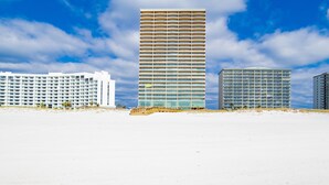 A beachfront hotel with multiple floors, a sandy beach in the foreground, and a clear blue sky with clouds. - New Complex!  Lazy River/Slide, 2BR Beachfront! (Orange Beach)