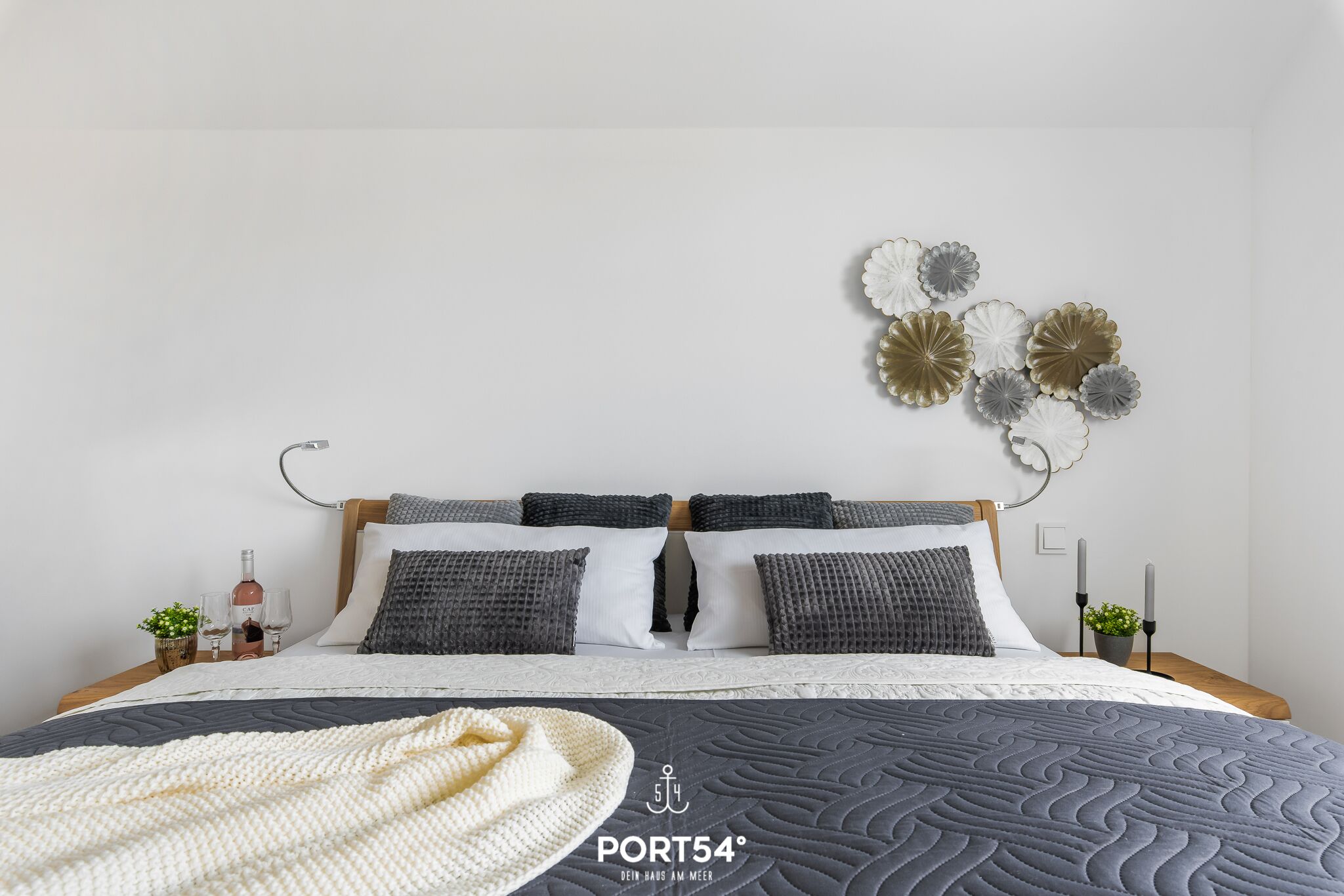 A neatly made bed with a grey quilt, white and grey pillows, and a white throw blanket. A decorative wall piece is above the bed.