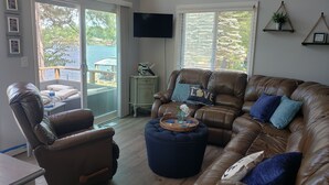 Ein Wohnzimmer mit einer Ledersofa, einem Hocker und einem Sessel. Ein Fernseher ist an der Wand montiert und es gibt ein Fenster mit Jalousien.