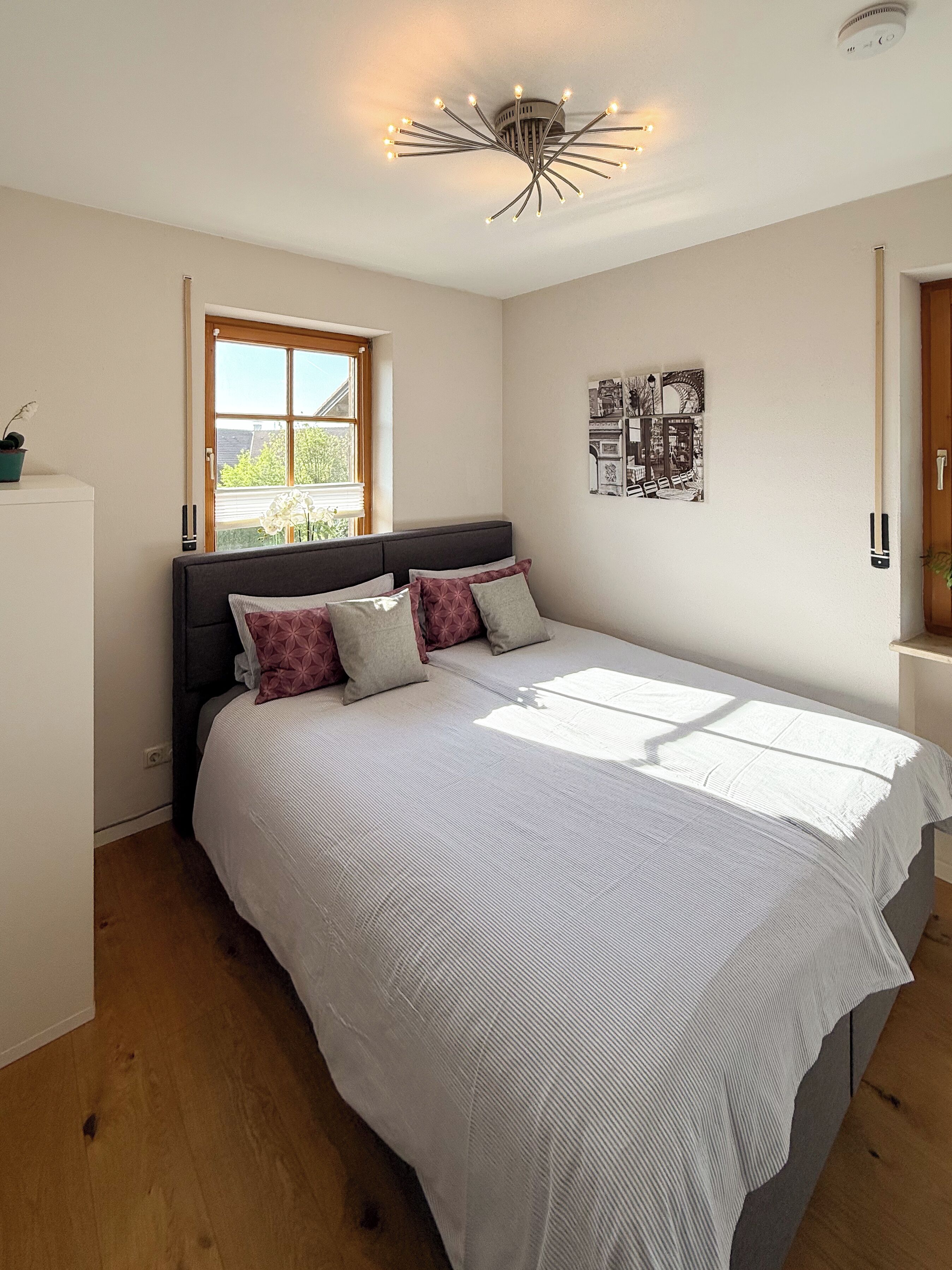 A bedroom with a large bed, a window with blinds, a ceiling light fixture, and a wall-mounted picture.