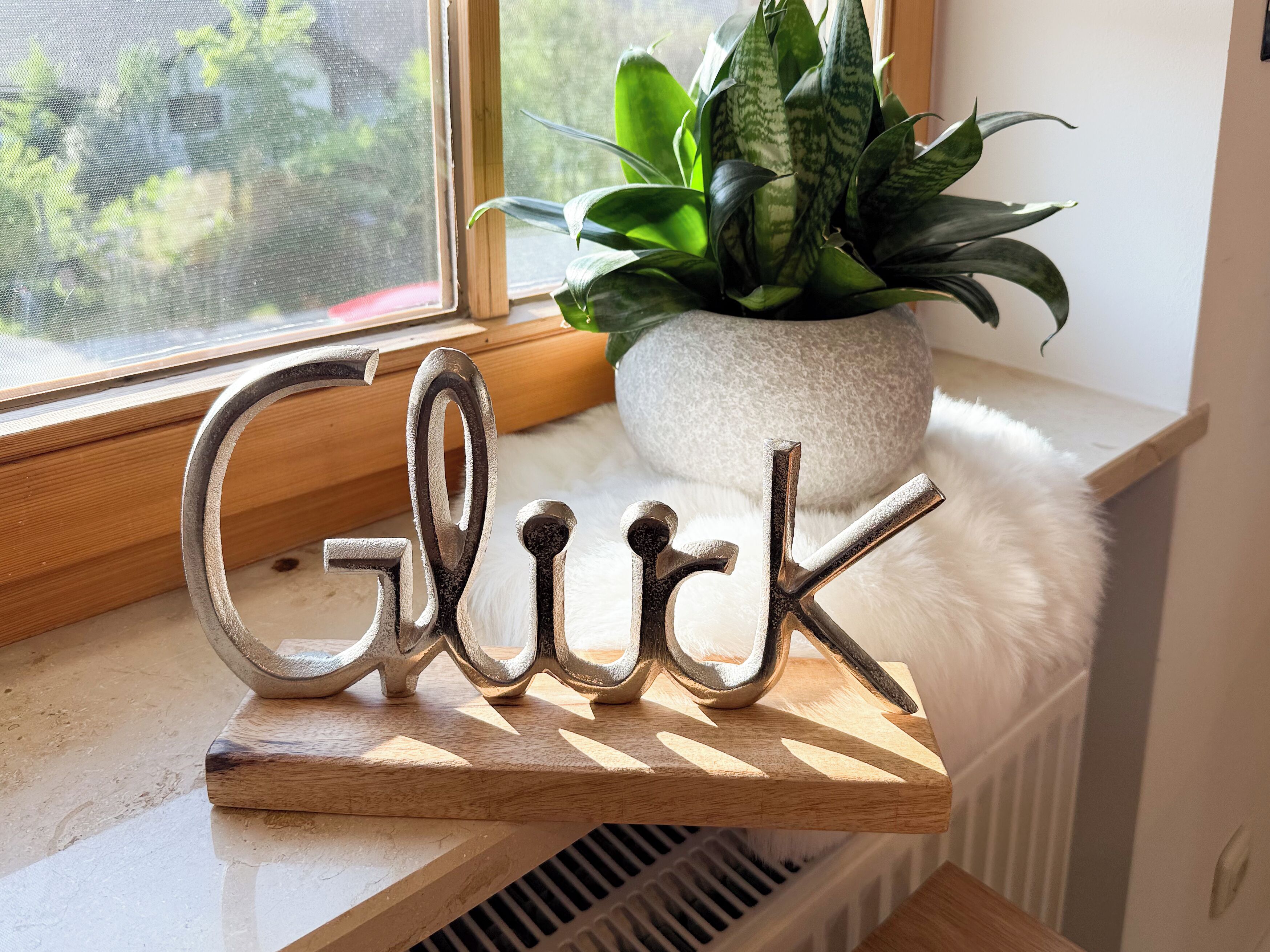 A decorative metal sign spelling "Glick" on a wooden shelf by a window with a plant and a stone pot.