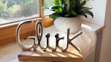 A decorative metal sign spelling "Glick" on a wooden shelf by a window with a plant and a stone pot.