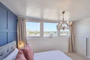 A bedroom with a large bed, a chandelier, a view of the beach, and a window with curtains. - Host & Stay | The Nest (Whitby)