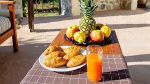 Ein Tisch mit einer Platte Croissants, einer Ananas und einem Glas Orangensaft, vor einer Steinwand mit einer Bank im Hintergrund.