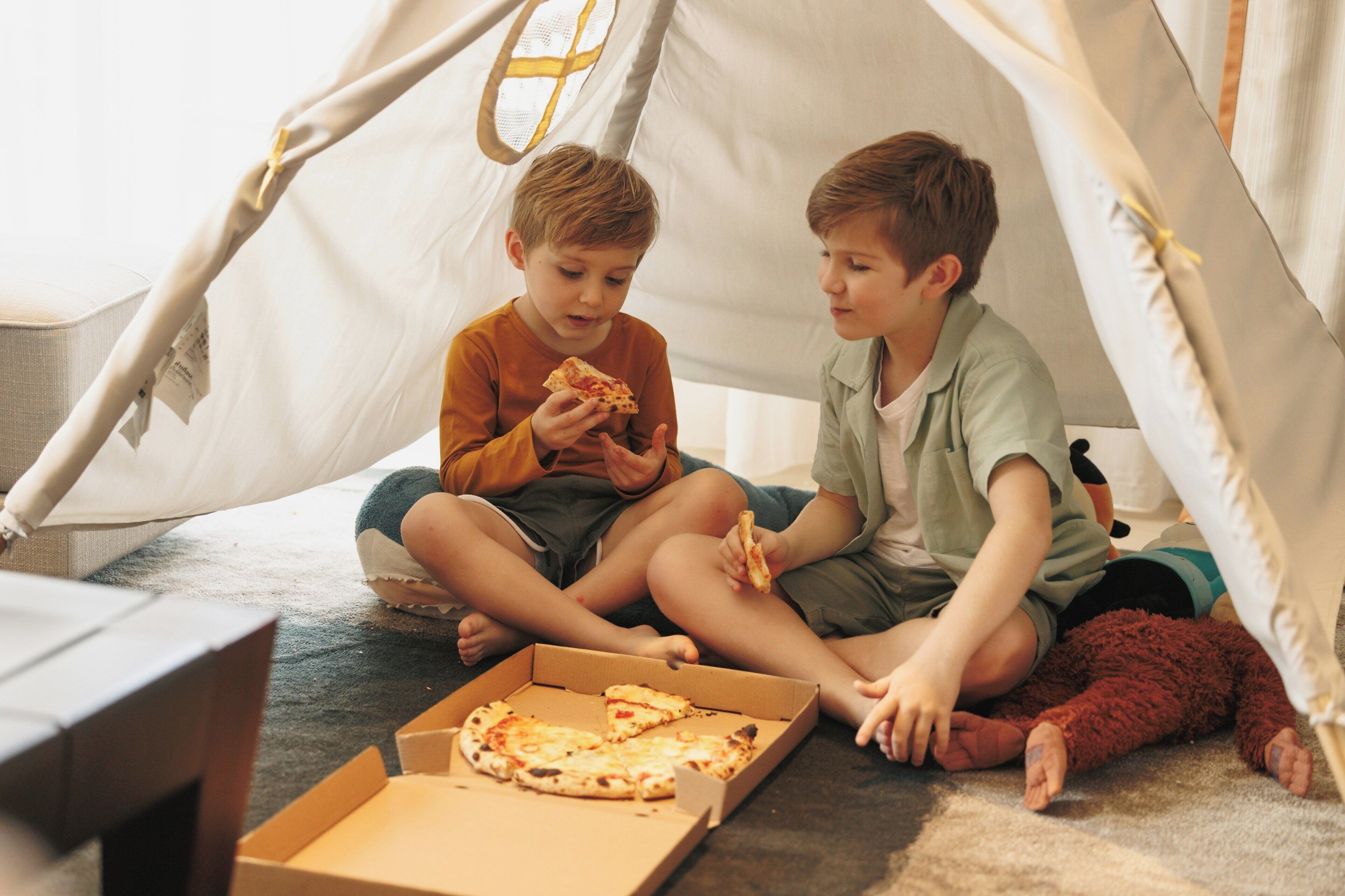 Two children sitting inside a tent-like structure with pizza boxes on the floor.