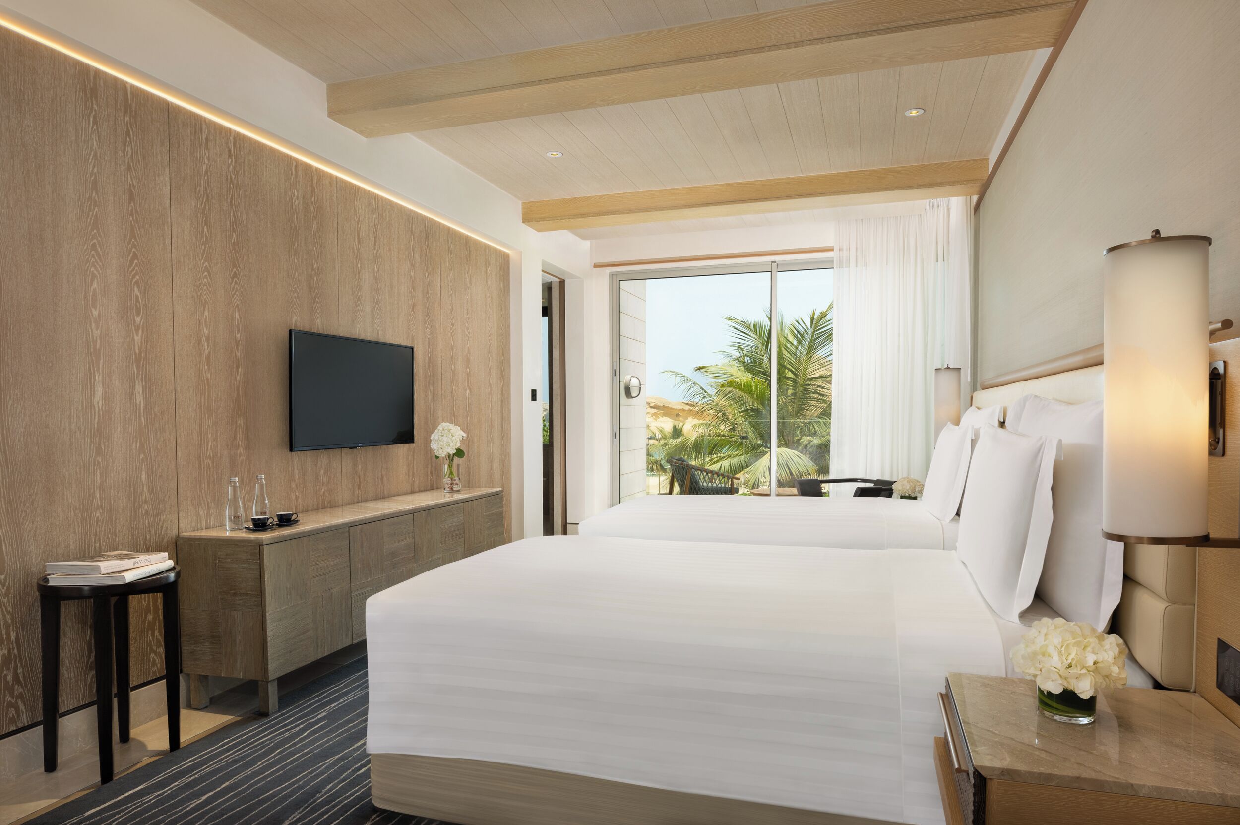 A modern hotel room with a large bed, a flat-screen TV, a wooden panel wall, a desk with a chair, and a view of palm trees through the window.