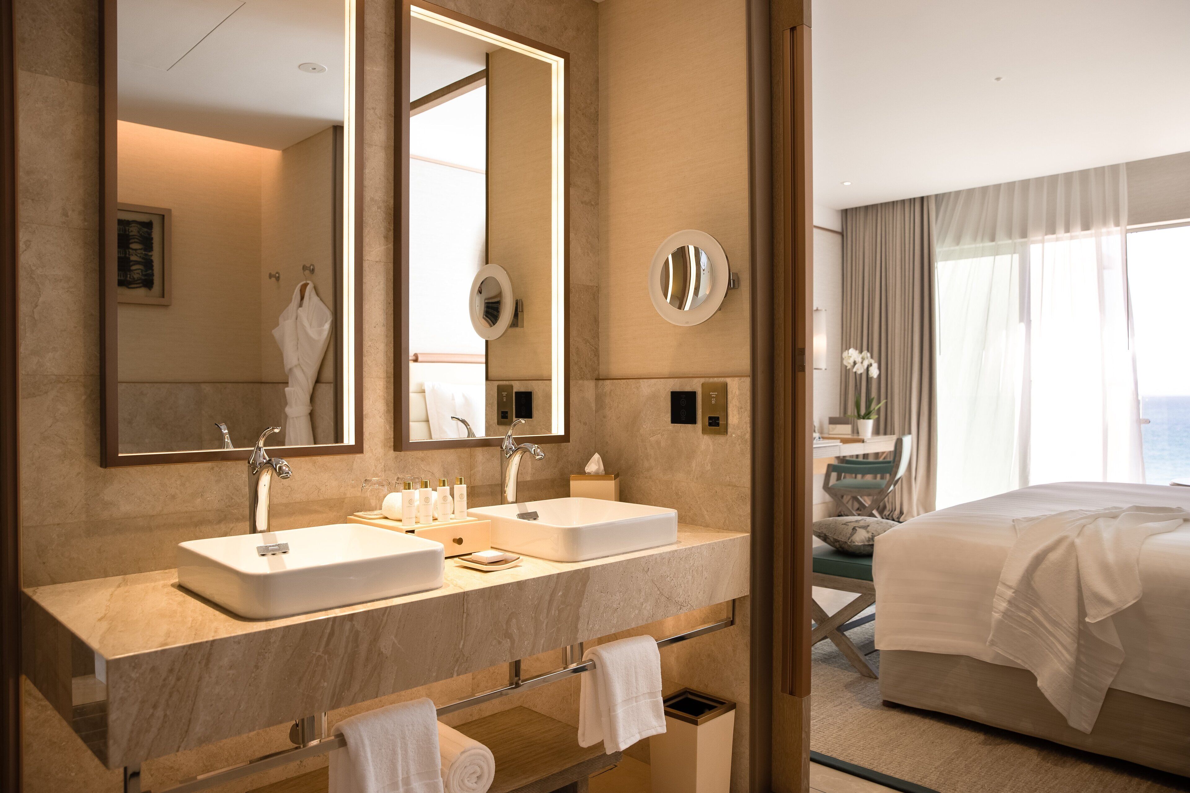 A hotel bathroom with two sinks, a large mirror, and a bed with a view of the sea.