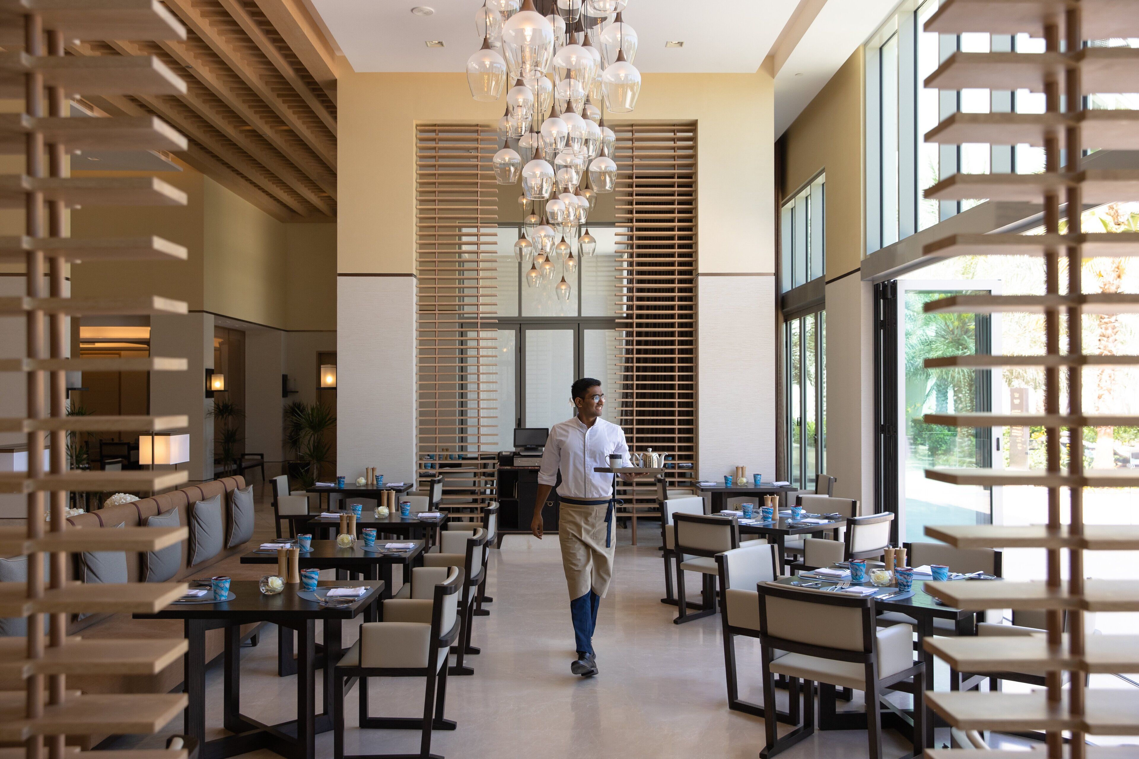 A spacious dining area with high ceilings, modern chandeliers, and large windows allowing natural light. Tables are neatly arranged with chairs, and a waiter is seen walking through the space.