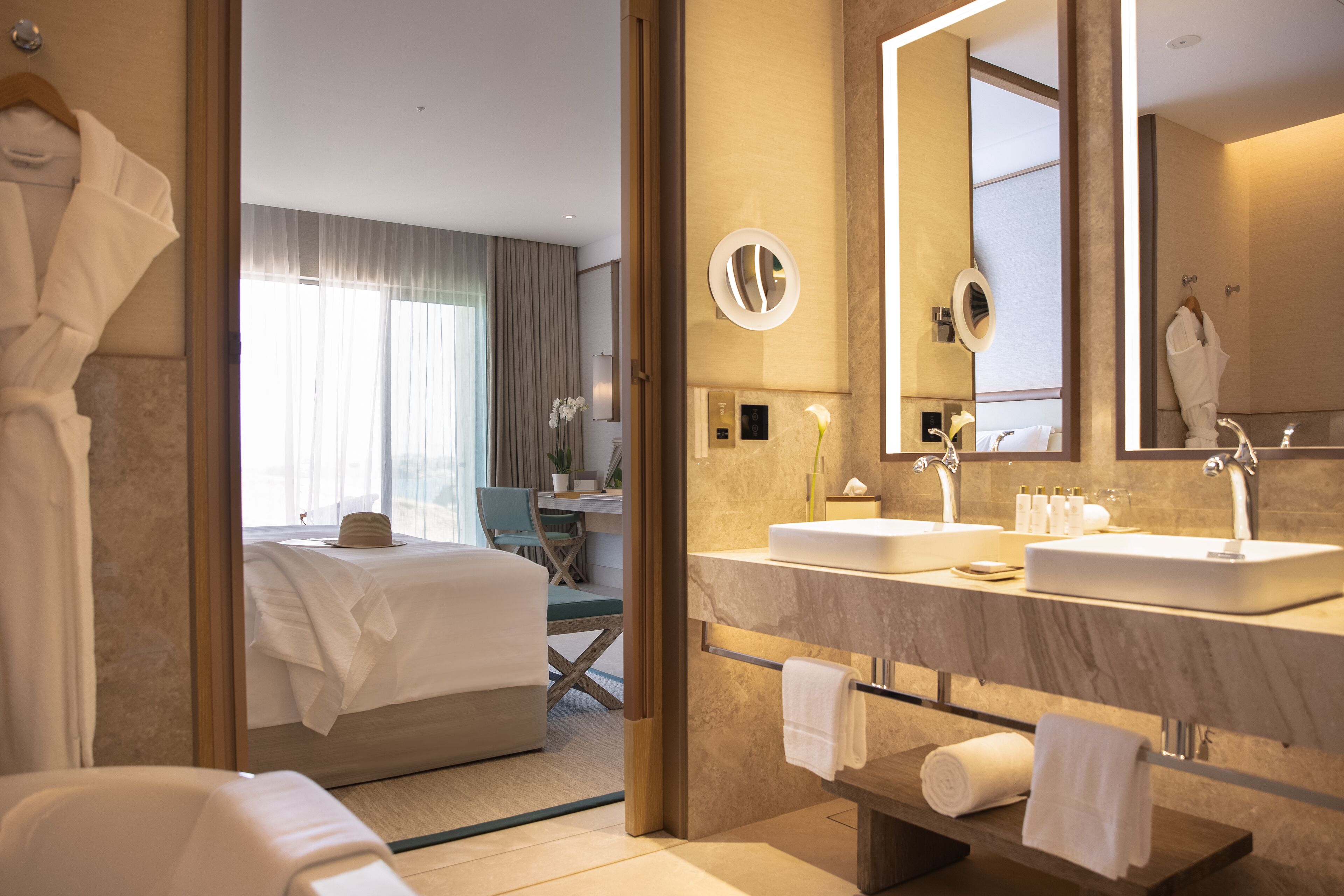 A hotel room with a large bed, a sitting area with a chair and table, a bathroom with a large mirror, two sinks, and towels.