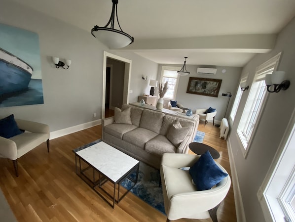 Ein großzügiges Wohnzimmer mit einem beigen Sofa, einem weißen Sessel und einem Couchtisch. An der Wand hängt ein Gemälde, und eine Chandelier sorgt für Beleuchtung. Ein Fenster lässt natürliches Licht herein.