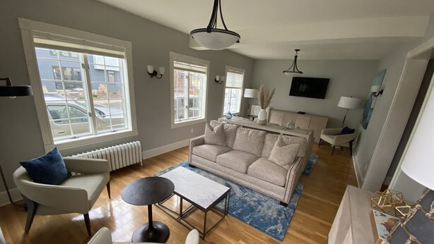 Un amplio salón con un sofá color beige, un sillón blanco y una mesita de centro. Amplias ventanas permiten la entrada de luz natural, y un televisor de pantalla plana está montado en la pared.