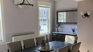 Un comedor con una mesa de madera oscura, sillas color beige, un radiador blanco, una ventana con persianas, un ventilador montado en la pared y un espejo con marco decorativo.