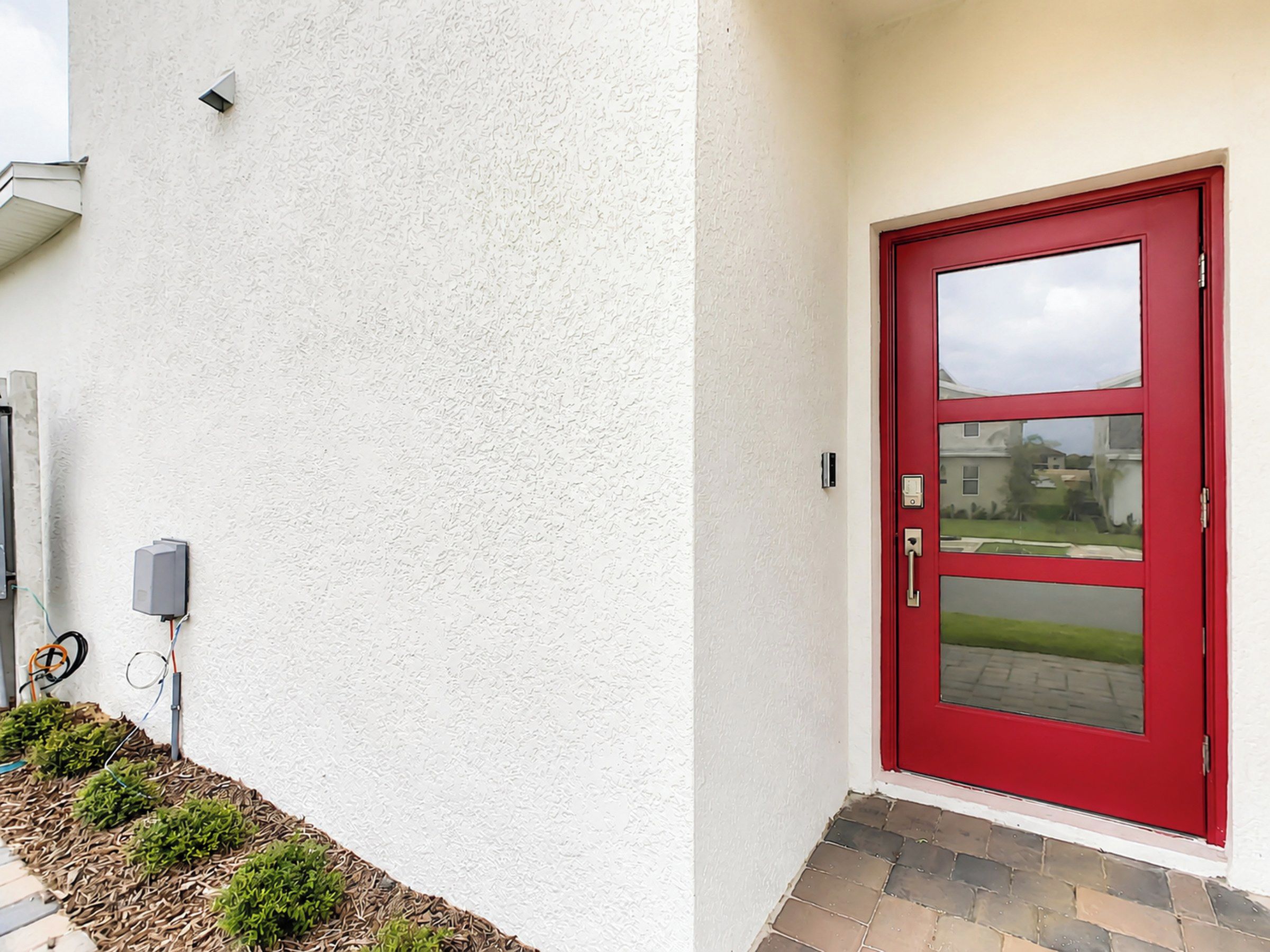Eine rote Tür mit Fensterglas, eine weiße, strukturierte Wand und ein kleiner Außenbereich mit Pflanzen und einem Pflasterweg.