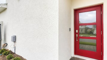 Eine rote Tür mit Fensterglas, eine weiße, strukturierte Wand und ein kleiner Außenbereich mit Pflanzen und einem Pflasterweg.