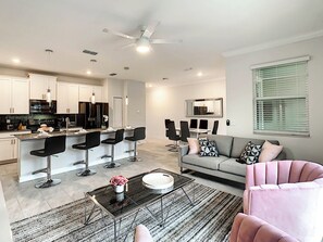 A modern kitchen with bar stools, a dining area with a sofa, and a ceiling fan.