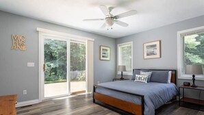 Ein Schlafzimmer mit Bett, Deckenventilator, großen Fenstern und Blick ins Freie.