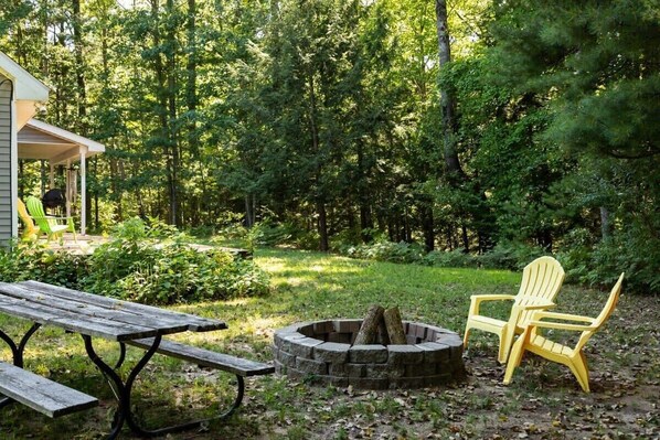 Un espacio de estar al aire libre rústico con una fogata, una mesa de madera y dos sillas Adirondack.