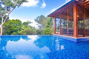 Ein klares, blaues Schwimmbecken mit einer Holzterrasse und einer Glaskonstruktion, umgeben von üppiger Vegetation.