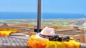 Una habitación con un ventanal grande que da a un paisaje costero, con una cama cubierta por un edredón amarillo y naranja, un sombrero blanco y unos binoculares.