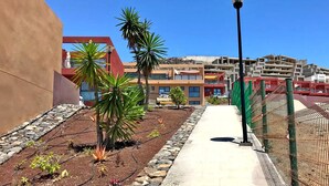 Un camino bordeado de palmeras y un farol, que conduce a un edificio de fachada roja y una zona de estacionamiento.