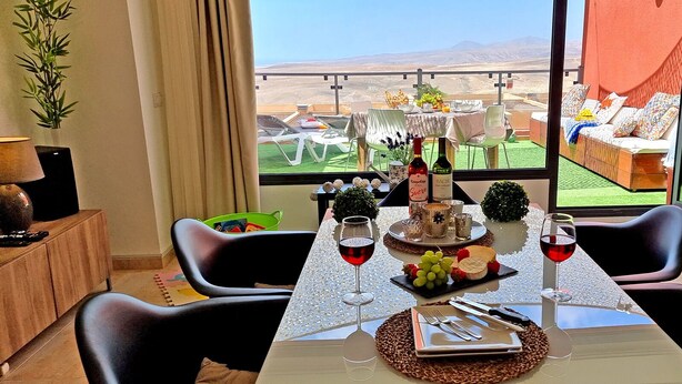 Una mesa de comedor dispuesta con copas de vino, una botella y una tabla de quesos, con vistas a un paisaje pintoresco.
