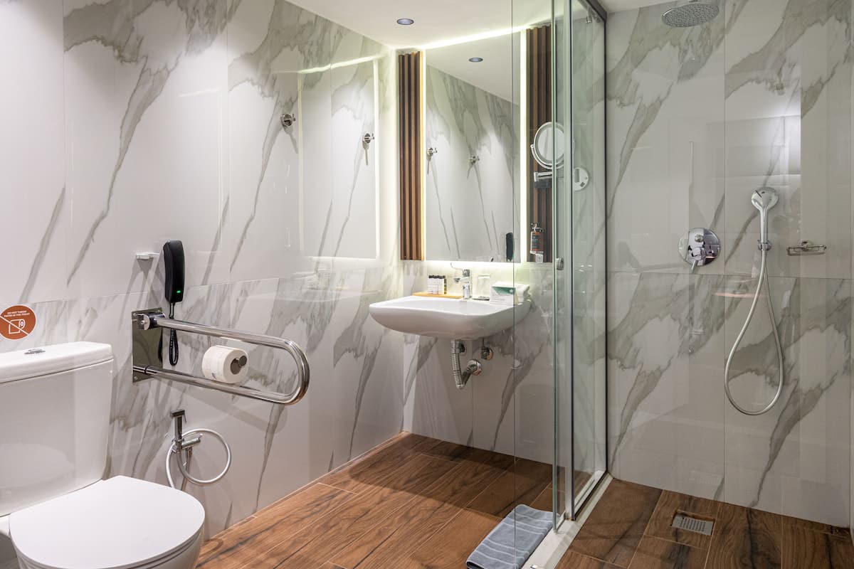 A modern bathroom with a marble wall, a wooden floor, a glass shower enclosure, and a toilet with a handrail.