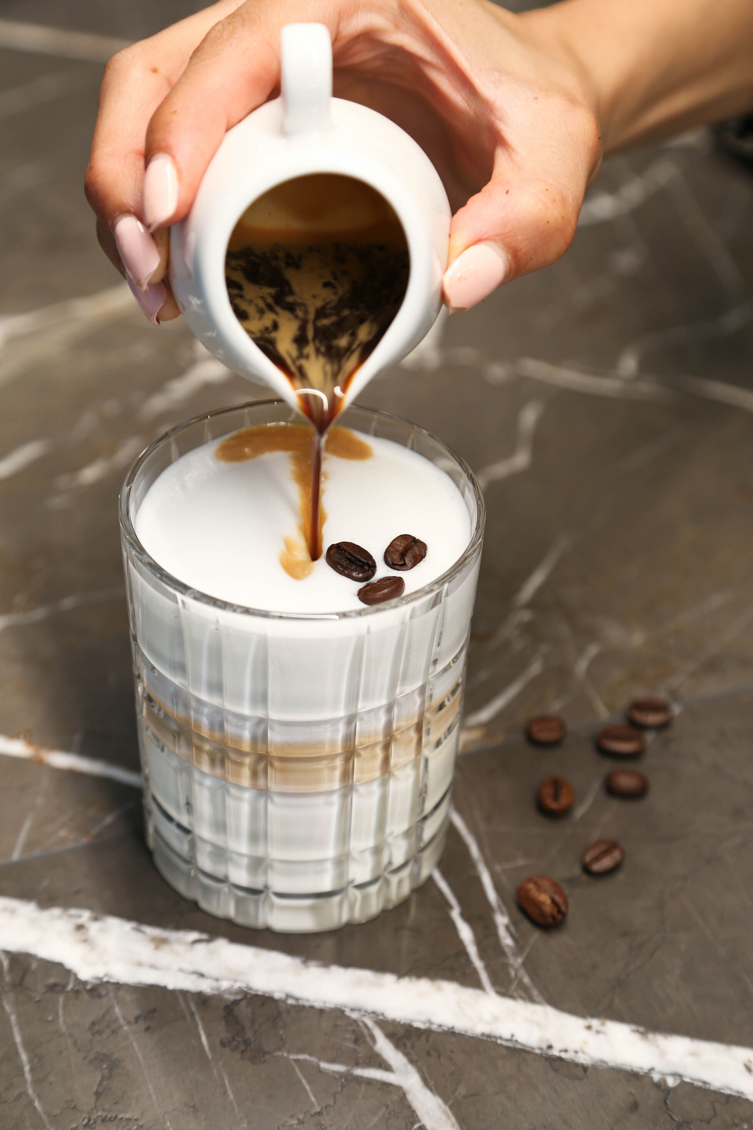 Una mano vertiendo un líquido oscuro en un vaso con base blanca, rodeado de granos de café sobre una superficie de mármol.