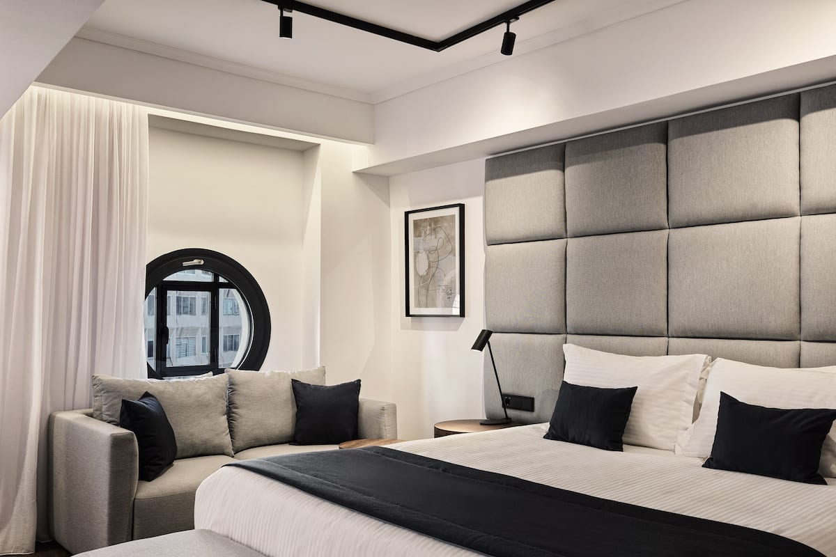 A modern bedroom with a large bed, a sofa, a round window, and a framed artwork on the wall.
