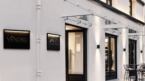 A modern building with a clean, white facade and black-framed windows. There are two signs on the wall: one for 'VANORO HOTEL' and another for 'ANZA LOUNGE BAR RESTAURANT'. - Vanoro Hotel (Thessaloniki)