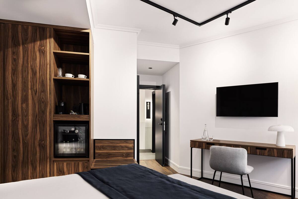 A modern hotel room with a bed, a wooden wardrobe, a desk with a chair, a television, and a minibar.
