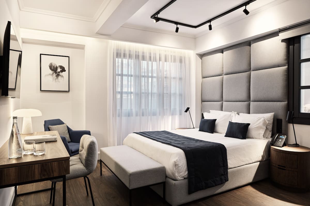 A modern hotel room with a large bed, a desk, a chair, and a bedside table.
