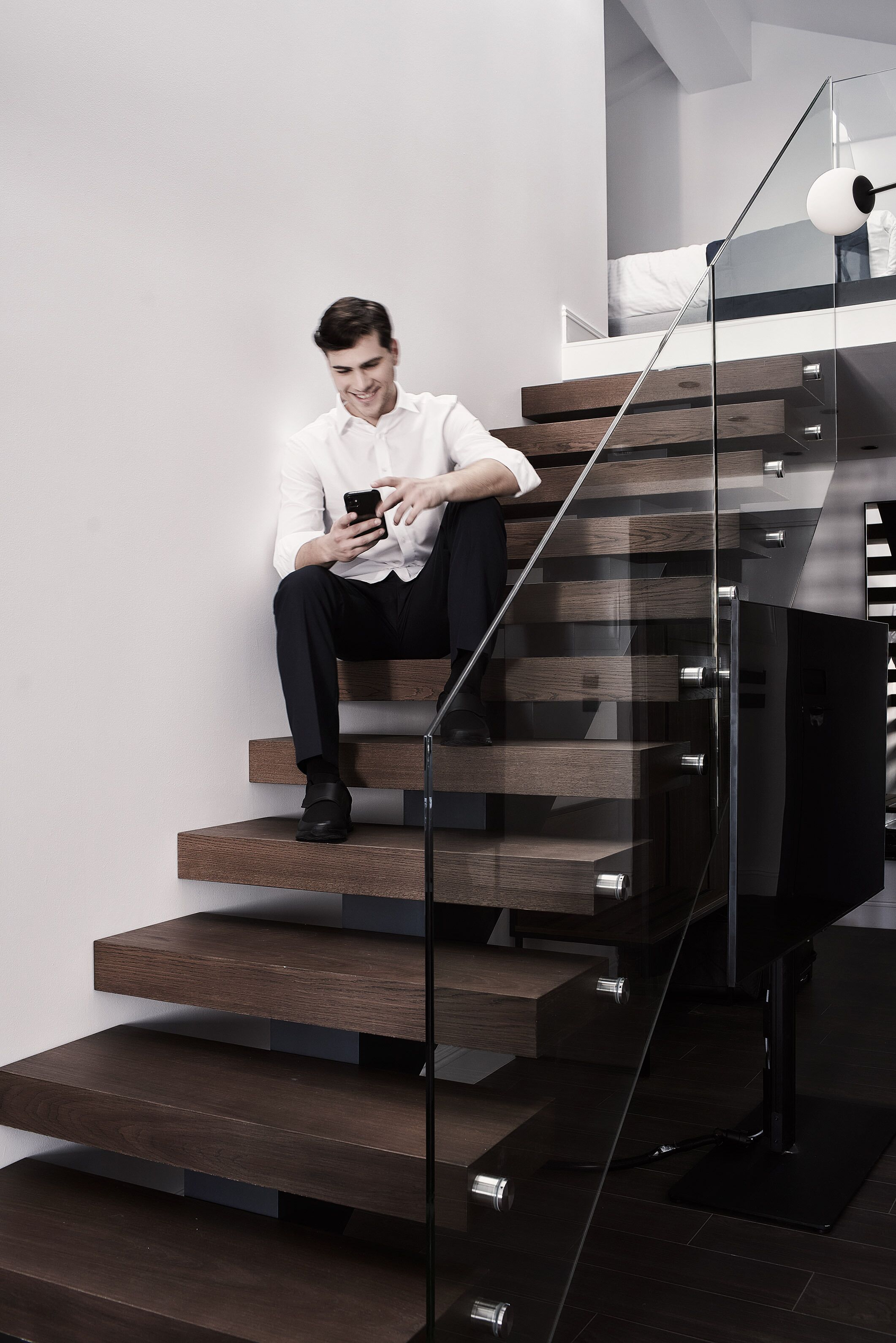 Un hombre sentado en una escalera moderna con barandillas de vidrio y escalones de madera.