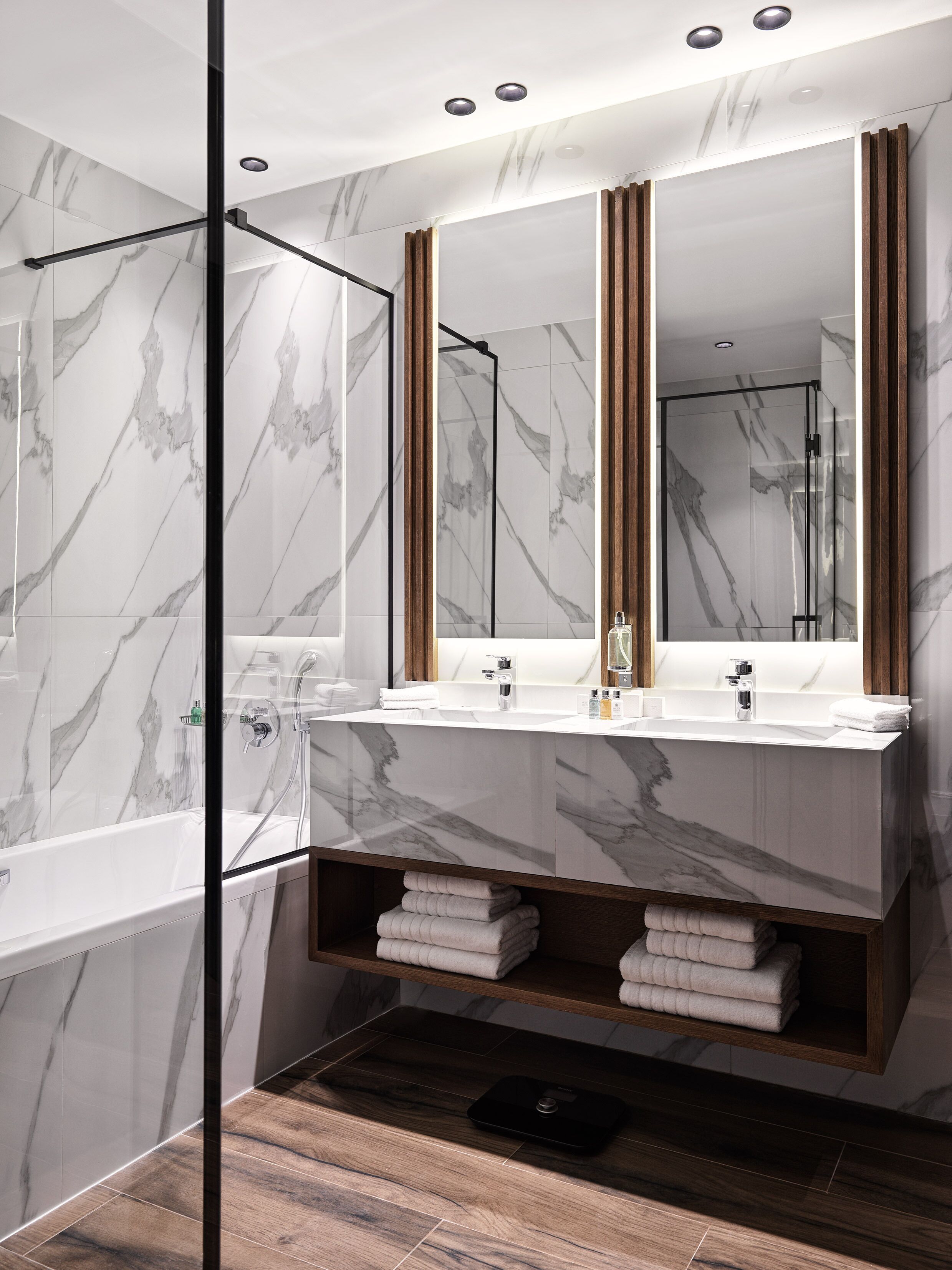 A modern bathroom with a large mirror, two sinks, a shower, and a wooden shelf with towels.