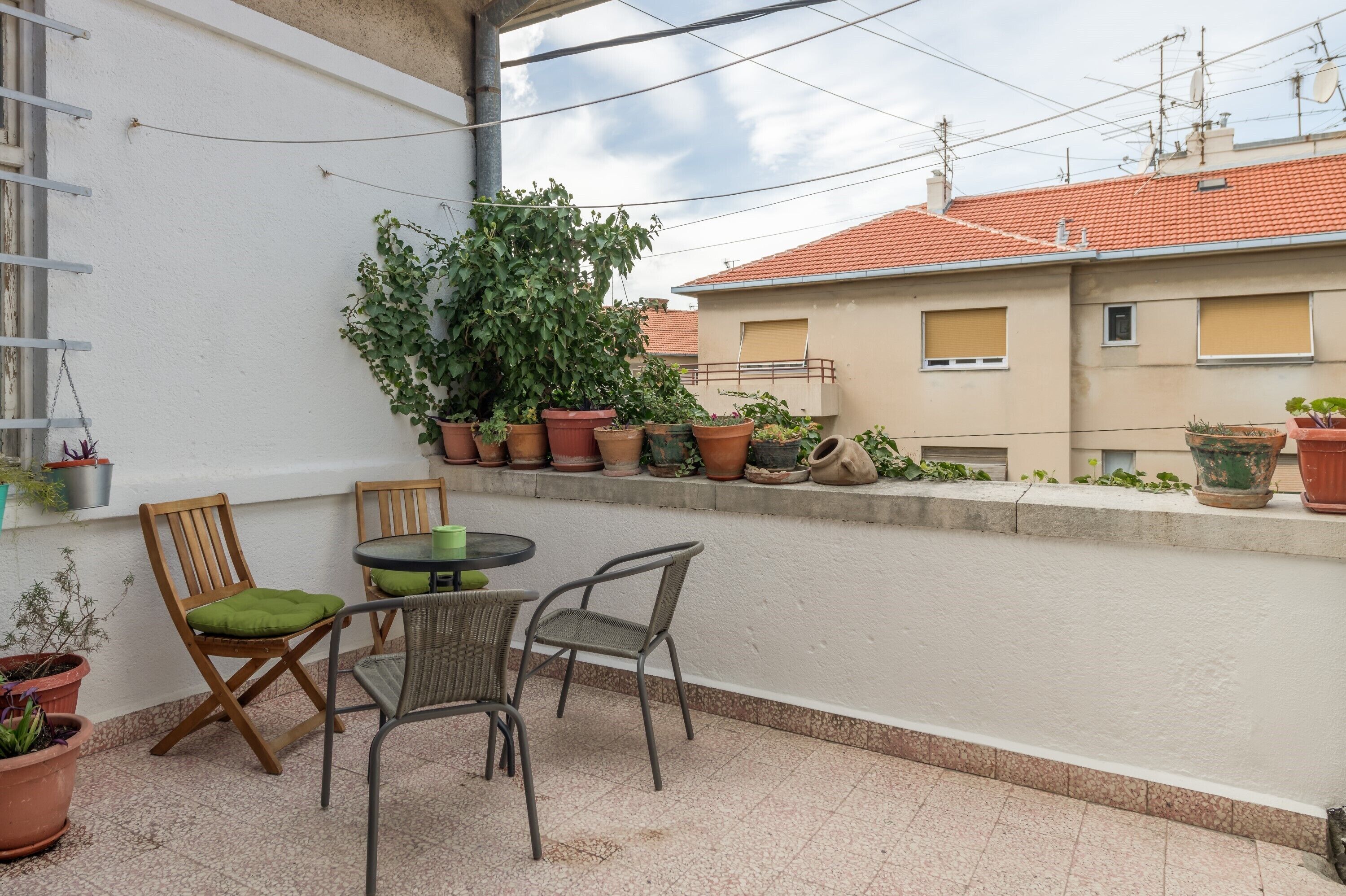 A balcony with potted plants, two wooden chairs, a table, and a view of neighboring buildings.