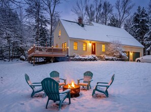 A house with a deck, a fire pit with chairs, and a snowy landscape.