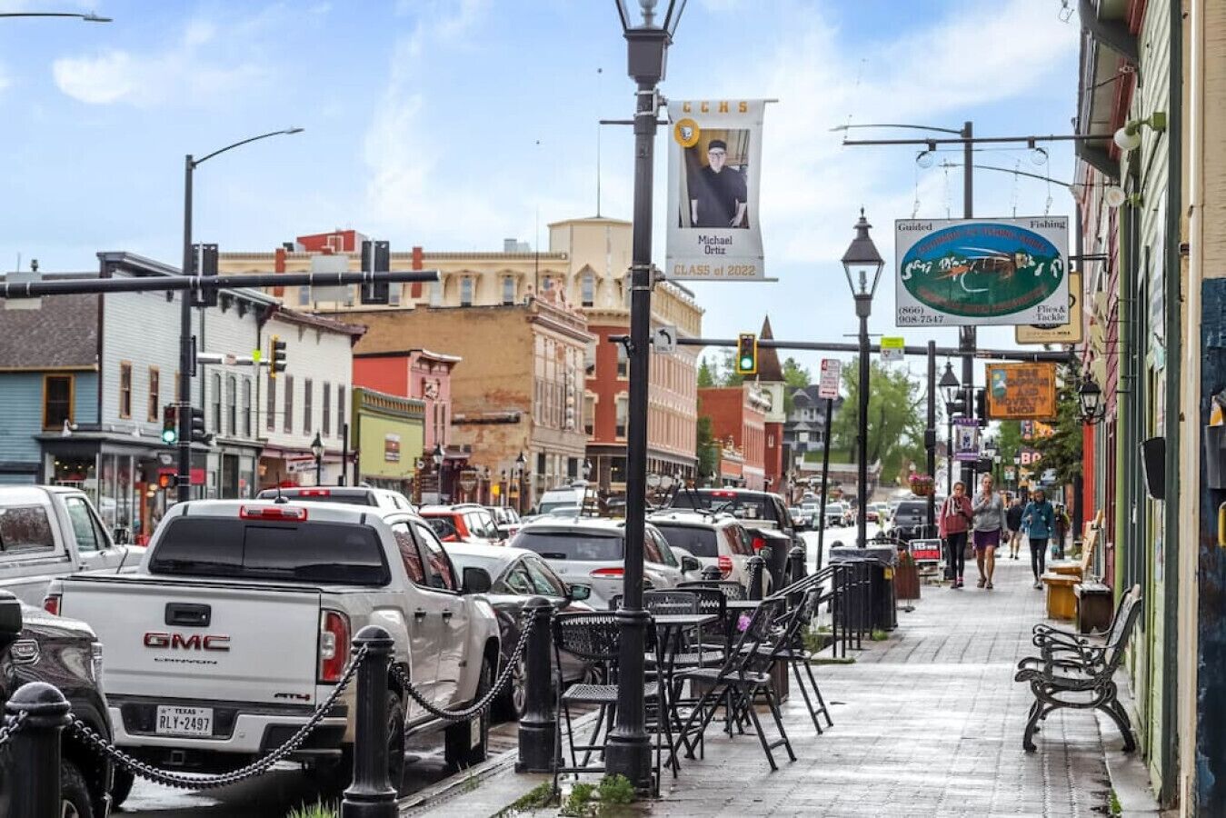 A busy street with parked vehicles, including a white GMC truck, and a sidewalk with outdoor seating.