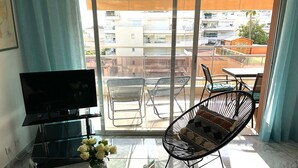 A living room with a TV, a glass coffee table, a chair, and a view of buildings through large windows. - Lovely and bright apartment on the fifth floor with a large terrace. Garage! (Cannes)