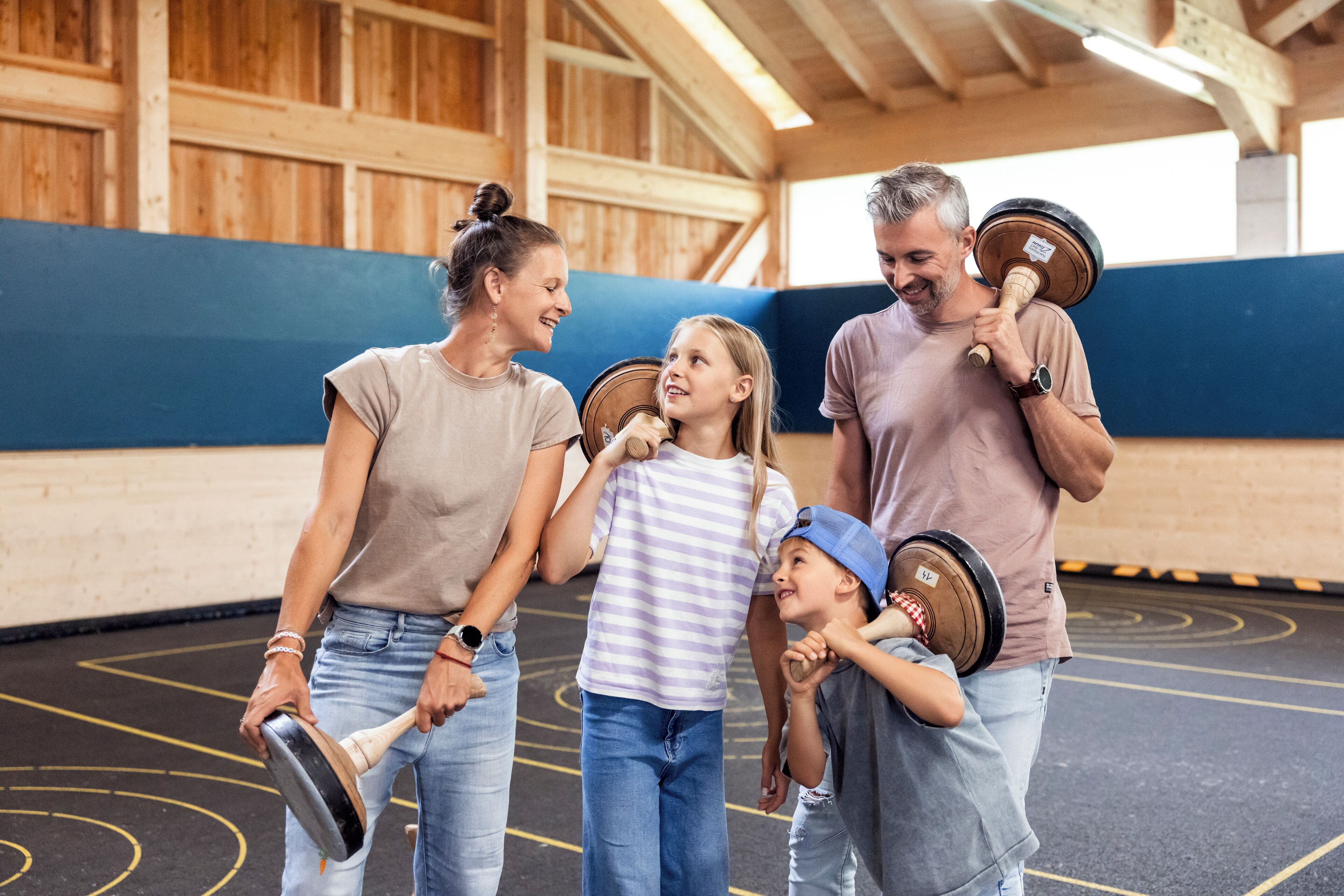 A family in an indoor gym with wooden walls and a blue floor, holding kettlebells.
