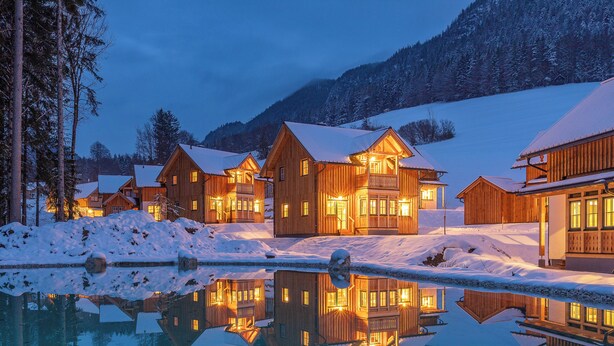 Una hilera de cabañas de madera con una piscina reflectante al frente, rodeadas de suelo y árboles cubiertos de nieve.