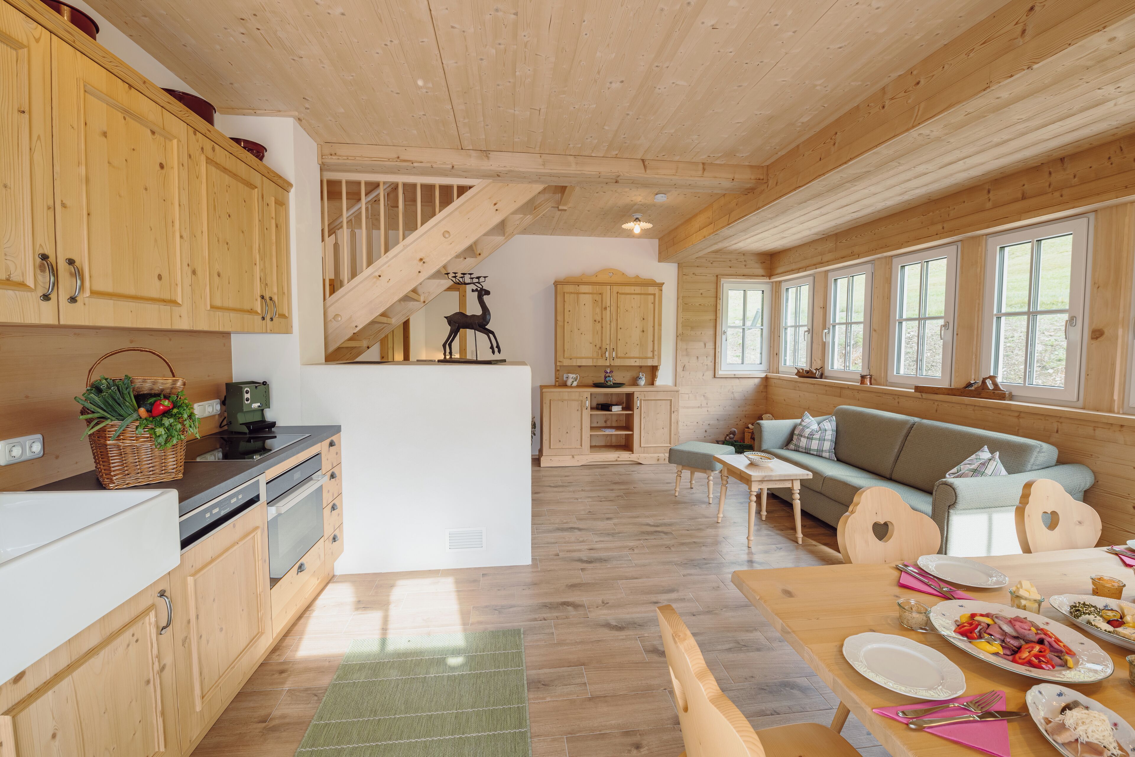 A cozy cabin interior with wooden floors, a dining area with a table set for a meal, a kitchen with wooden cabinets, and a staircase leading to an upper level.