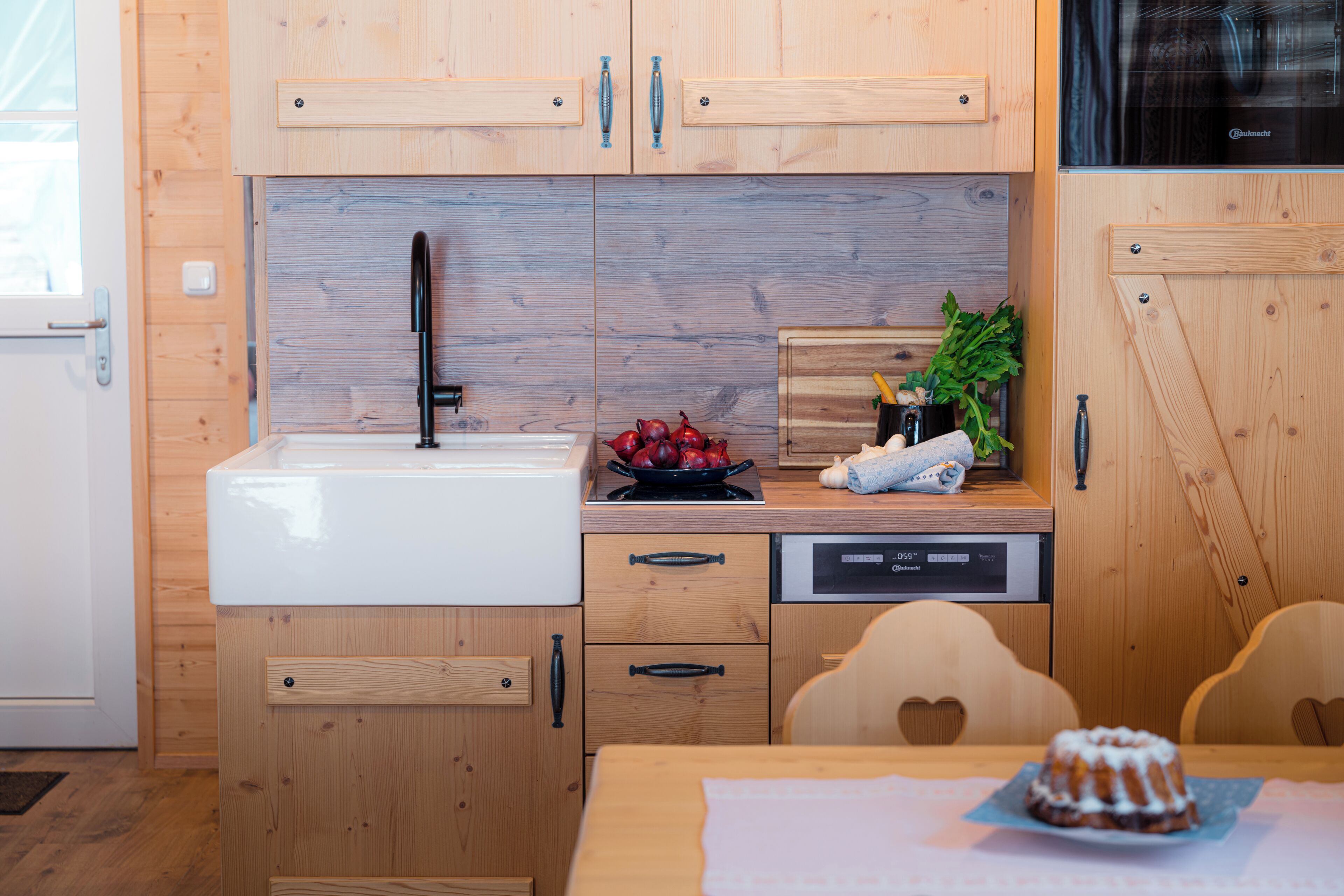 A compact kitchen with wooden cabinetry, a white sink, a black faucet, and a countertop with a bowl of fruit and a plant.